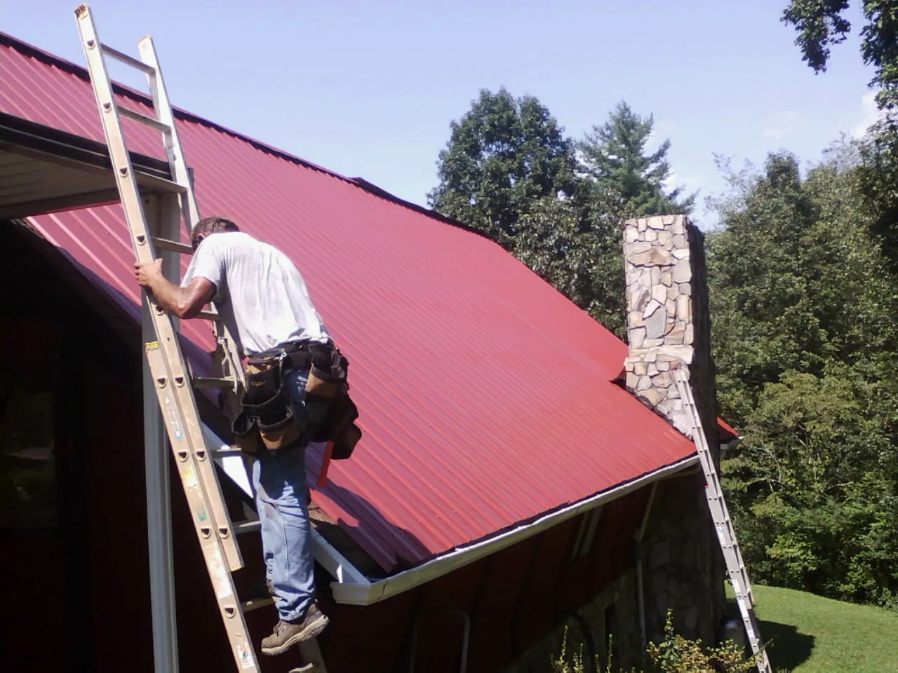 Roofer on a ladder beside a red metal roof and stone chimney — Soffit & Fascia Repair by Roofing Brothers in Rio Medina